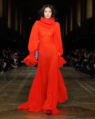 A model wearing a red sheer silk gown with ruffled sleeves and a ruff while walking at the F/W 25 Alexander McQueen show.