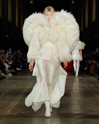 A model wearing a cream silk-and-shearling bolero over a sheer silk gown with white boots at the Alexander McQueen F/W 25 show during Paris Fashion Week.