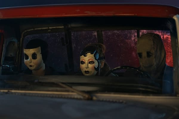 Three figures wearing creepy masks sit in a truck's front seat, creating a tense atmosphere. This still is from 'The Strangers - Chapter 3,' with a backdrop featuring ominous colors.