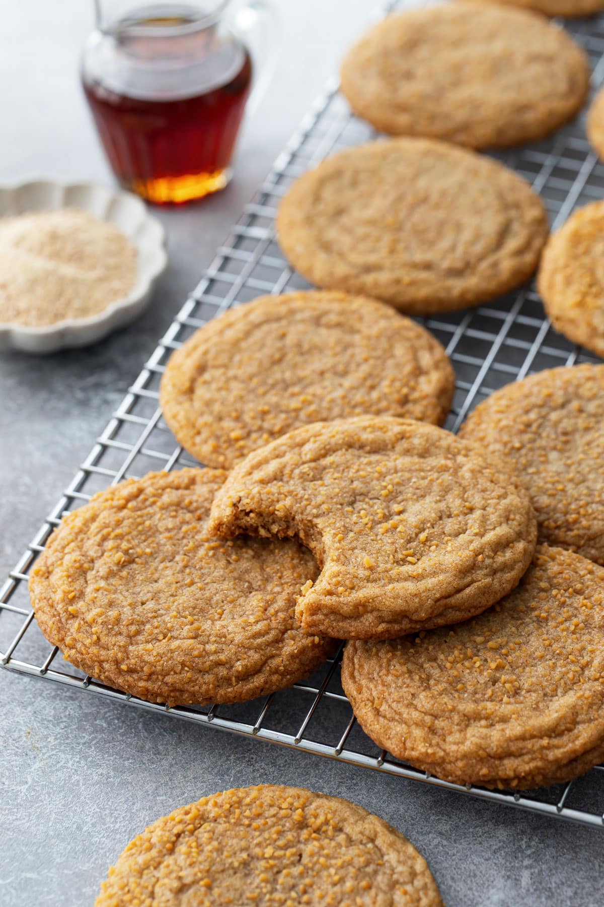 Chewy Maple Brown Butter Sugar Cookies on a wire baking rack, with a small pitcher of maple syrup and a bowl of maple sugar in the background.