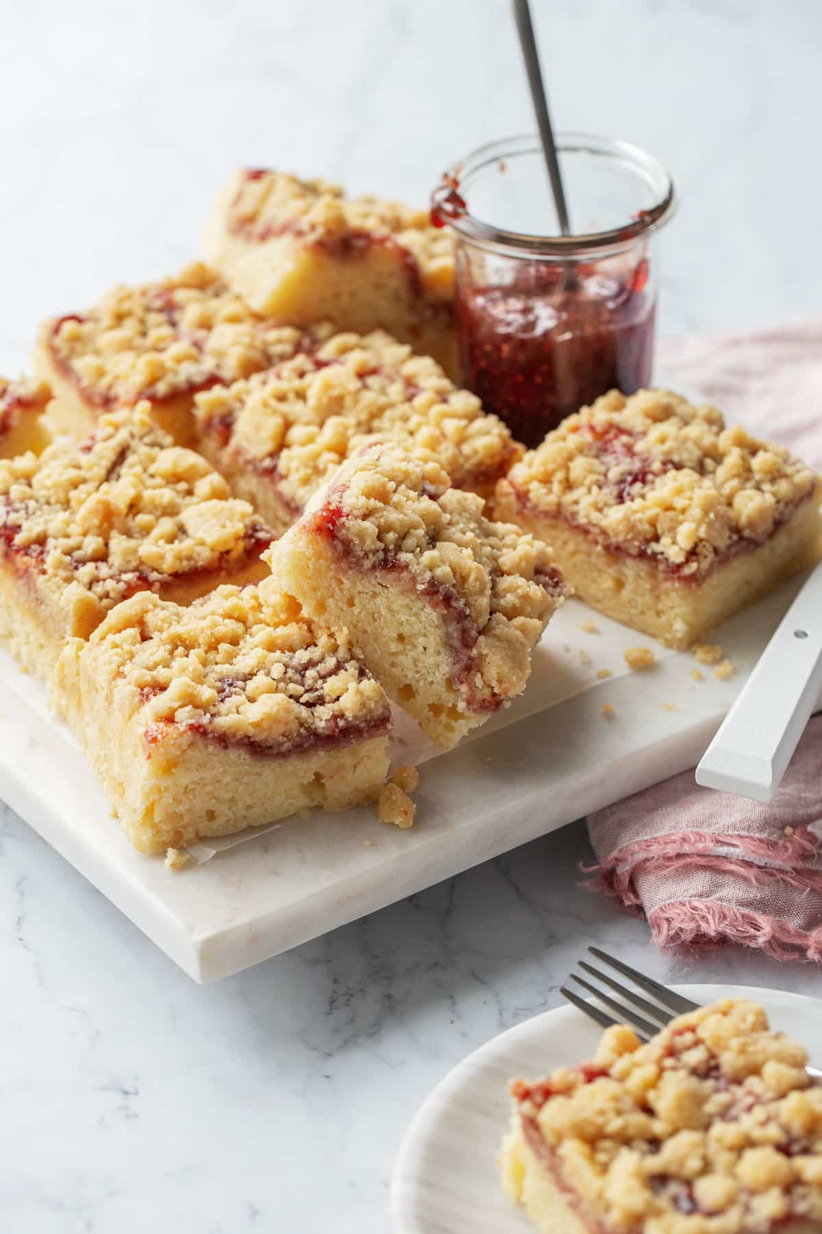 Squares of Sour Cream Crumb Cake with Jam Swirl and crumb topping on a marble serving plate with a jar of raspberry jam.