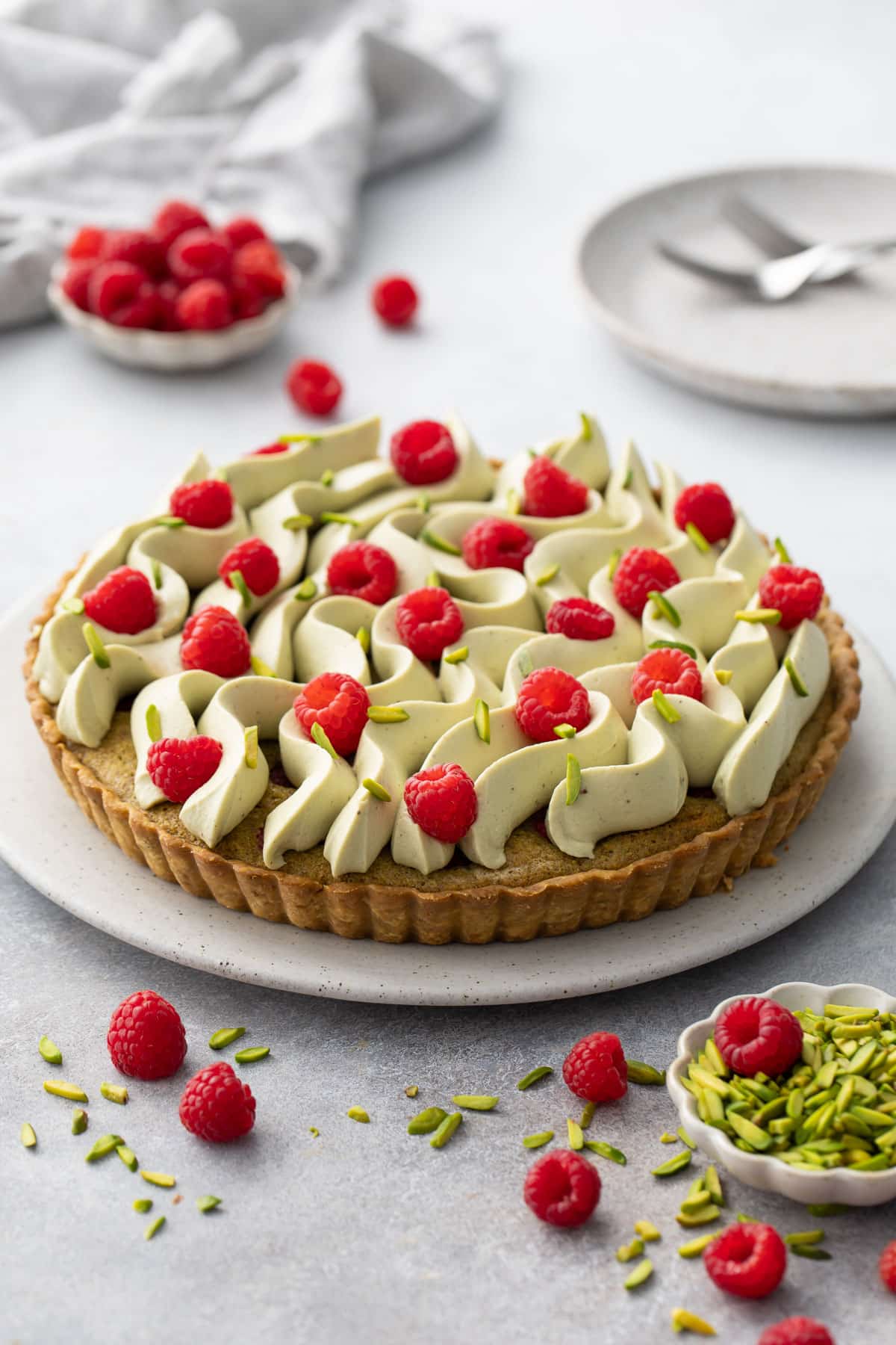 Raspberry & Pistachio Frangipane Tart with Whipped Pistachio Ganache piped in waves on a flat cake plate with rasberries and slivered pistachios scattered on top and around.