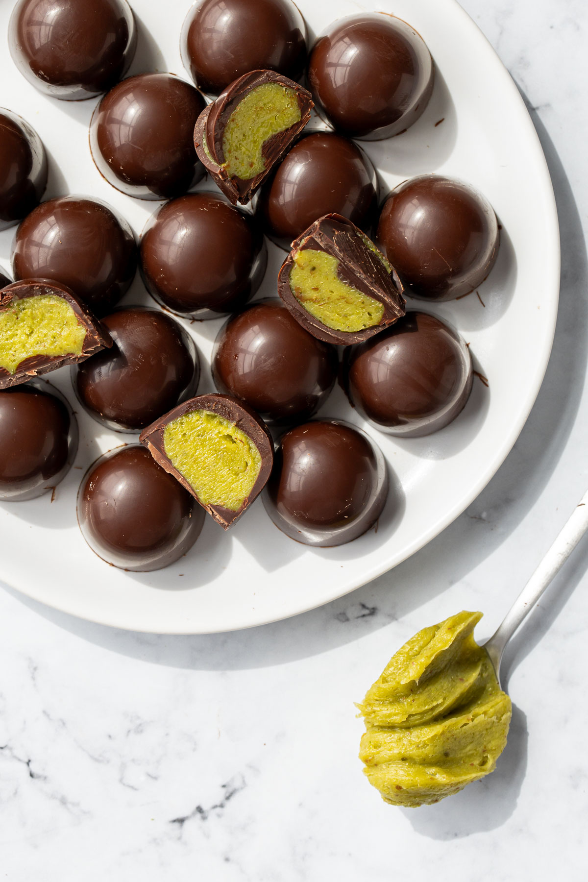 Plate piled with dark chocolate truffles, a few cut in half to show the bright green homemade pistachio ganache inside, with a spoon of ganache on the side.