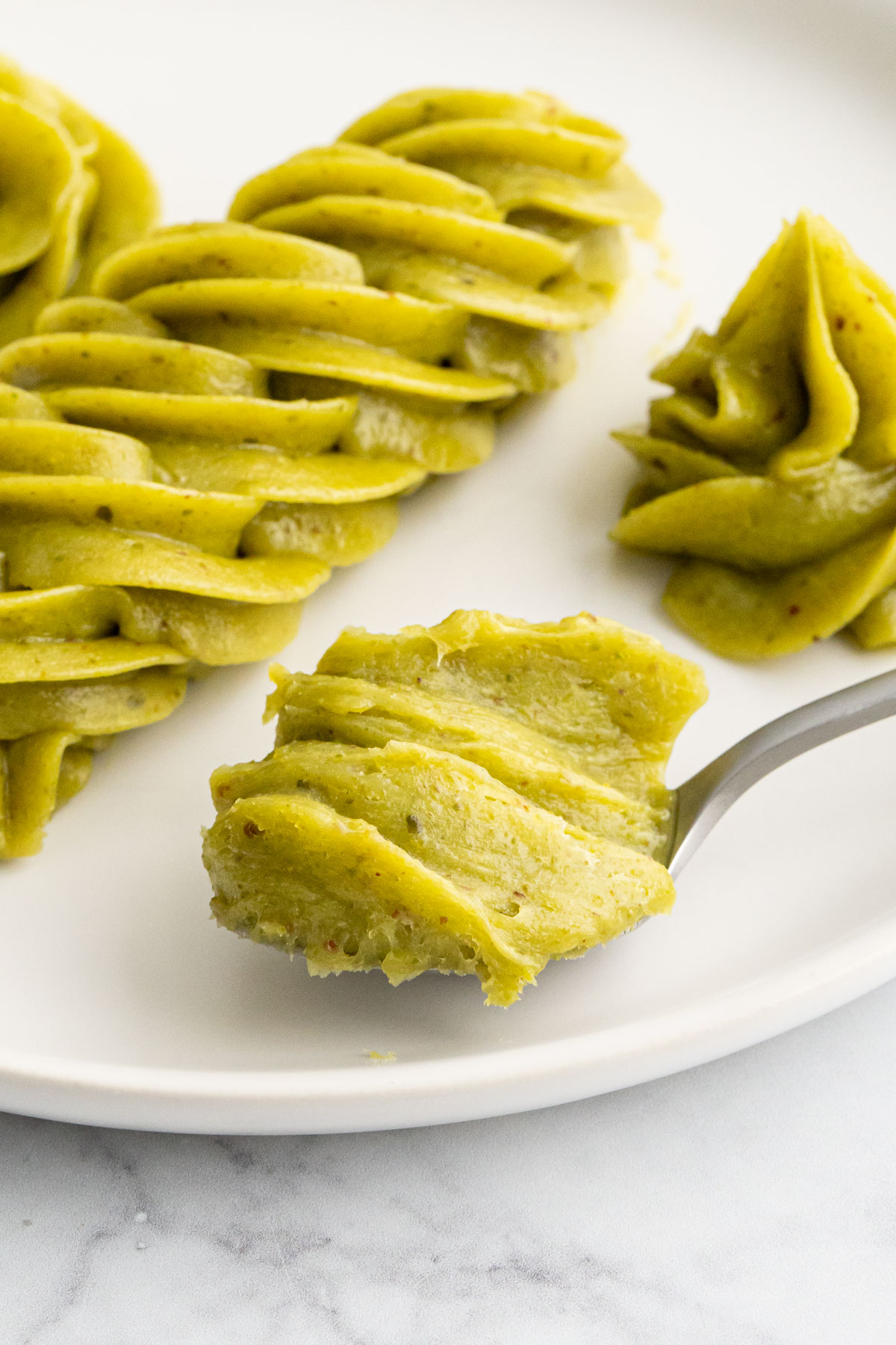 Spoonful of bright green pistachio white chocolate ganache on a plate along with some piped shapes.