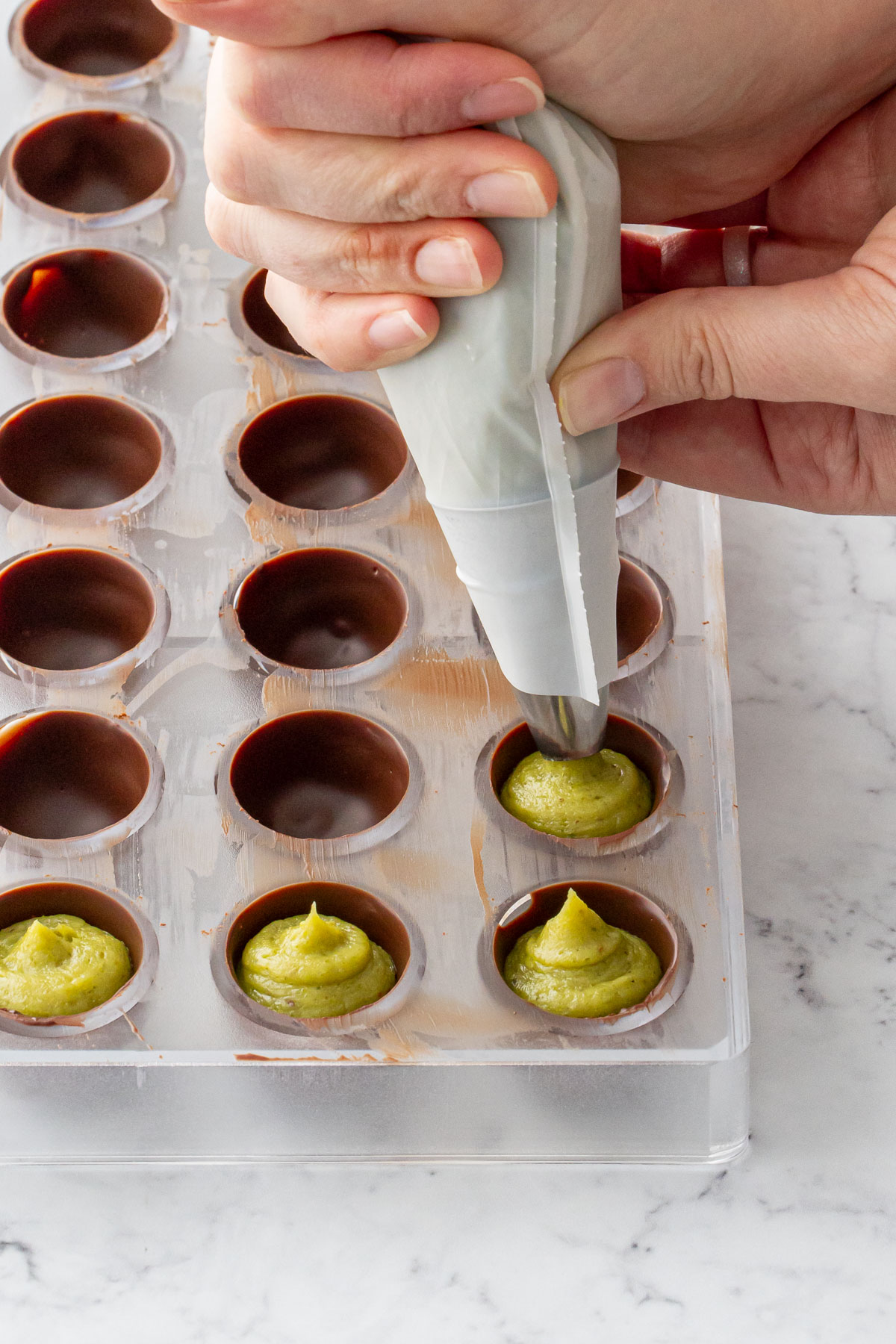 Piping bright green pistachio ganache into dark chocolate truffles in a polycarbonate mold.