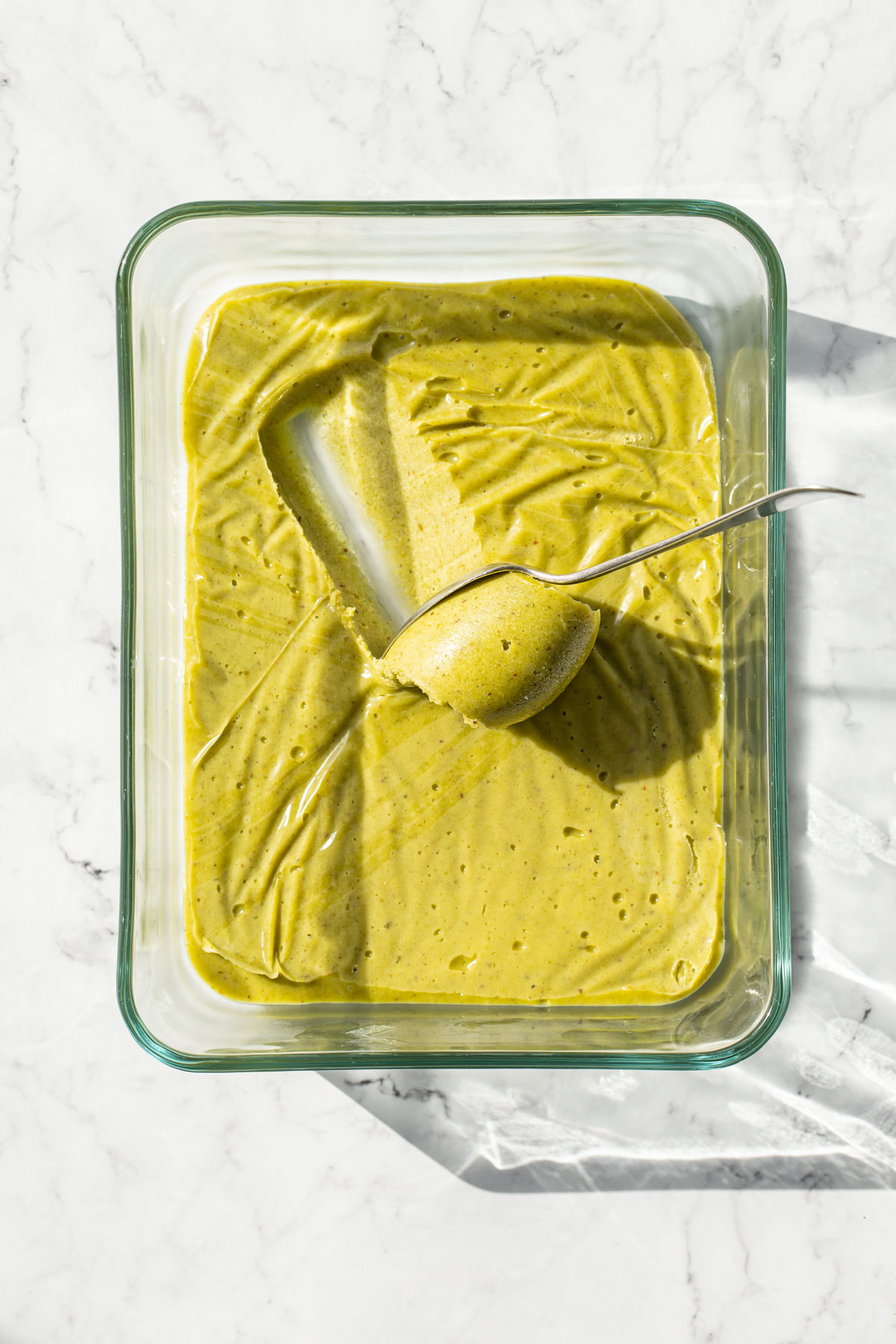 Overhead view, shallow glass dish with chilled pistachio ganache and a spoon scooping a curl of ganache.