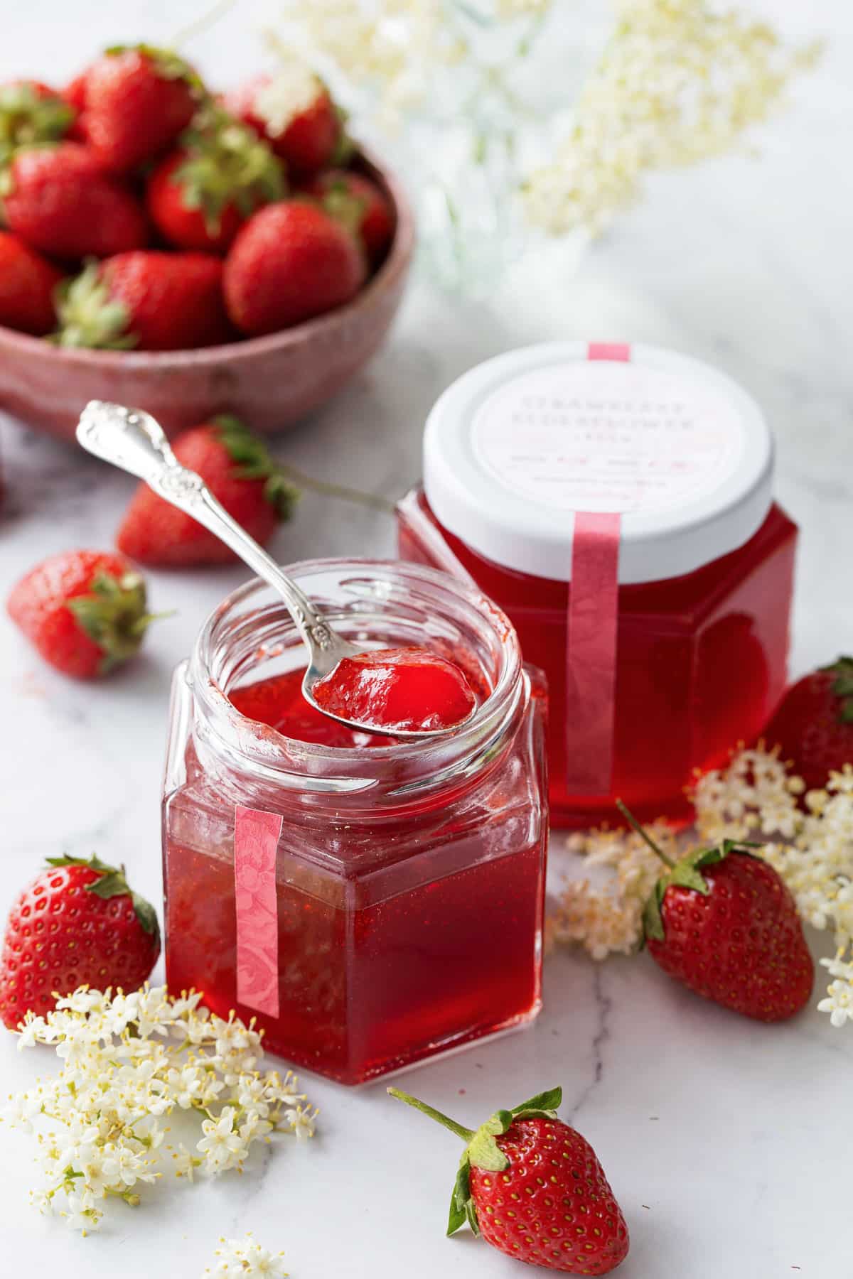 Two jars of Strawberry Elderflower Jelly, one open with a spoonful sitting on top to show the jelly texture, with fresh strawberries and elderflowers scattered around.