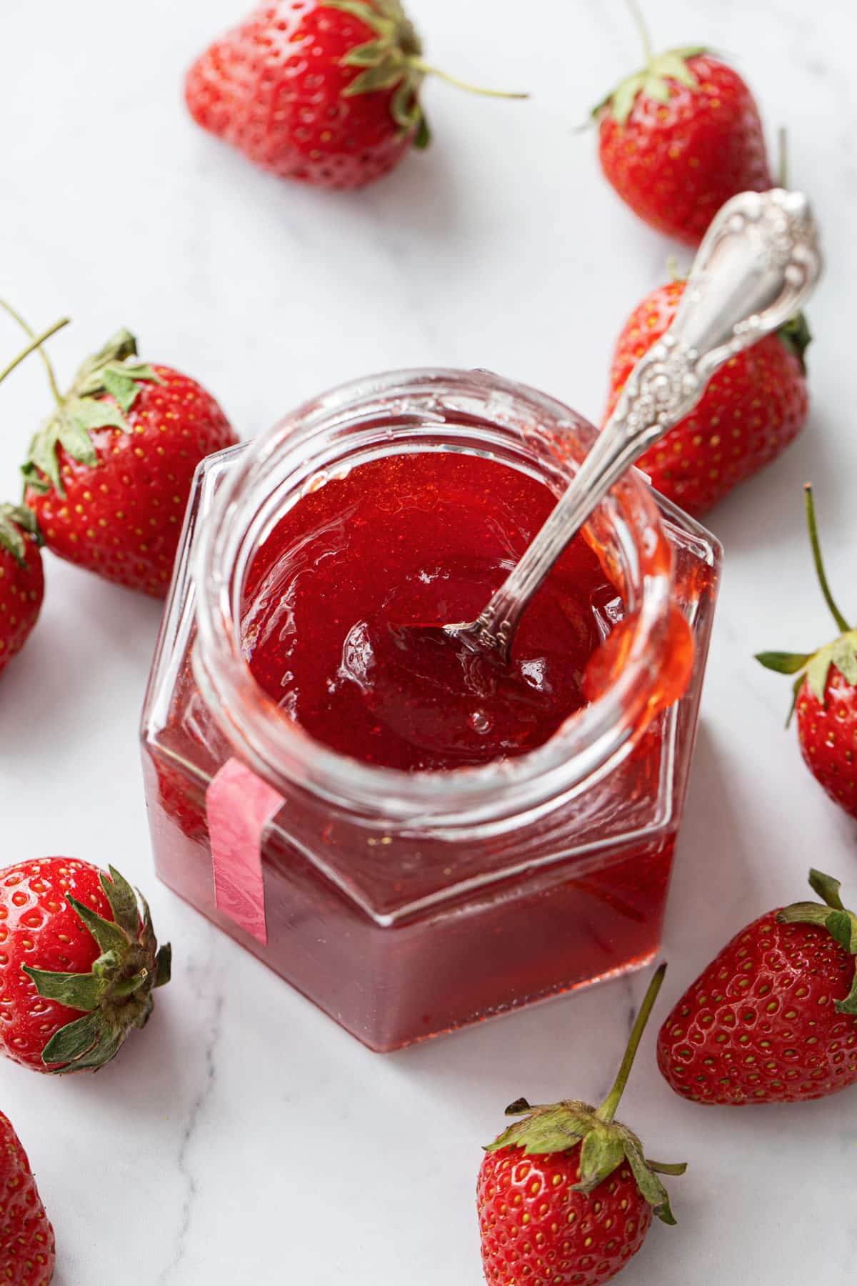 Open jar of Strawberry Elderflower Jelly with a silver spoon inside it, showing the jelly-like texture, with a few berries scattered around.