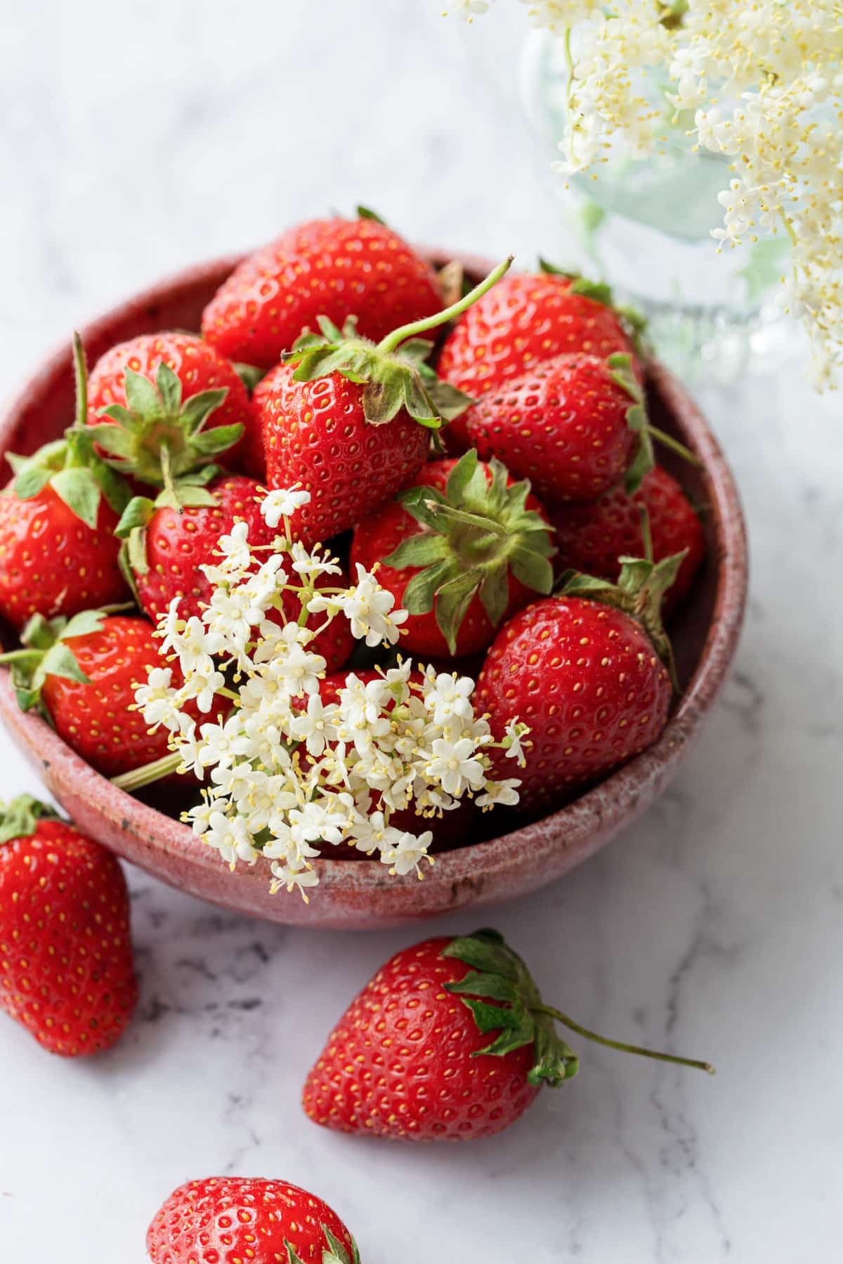 Bowl of fresh strawberries and sprigs of elder flowers on a marble background.