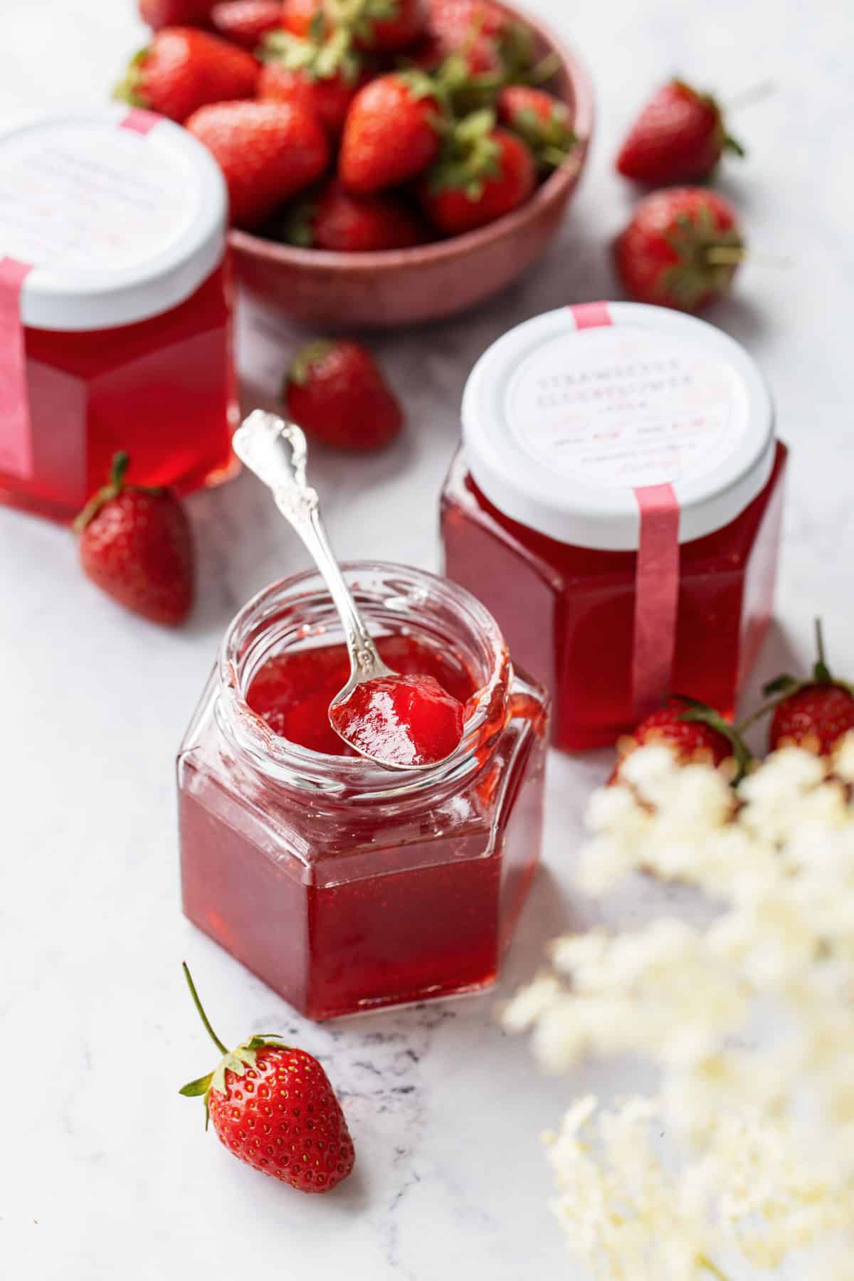 Two jars of Strawberry Elderflower Jelly, one open with a spoonful sitting on top to show the jelly texture, with fresh strawberries and out of focus elderflowers in the foreground.