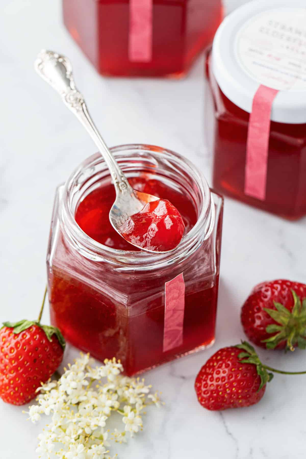 Open jar of Strawberry Elderflower Jelly with a spoonful resting on the rim, and a few berries and flowers scattered around.