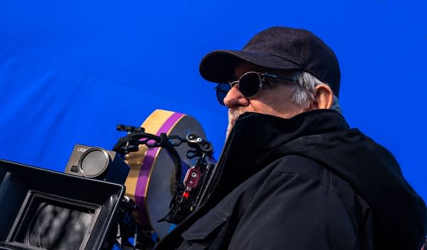 Director Steven Spielberg on the set of 'Disclosure Day,' observing through sunglasses, with a camera equipment setup in front against a vibrant blue background.