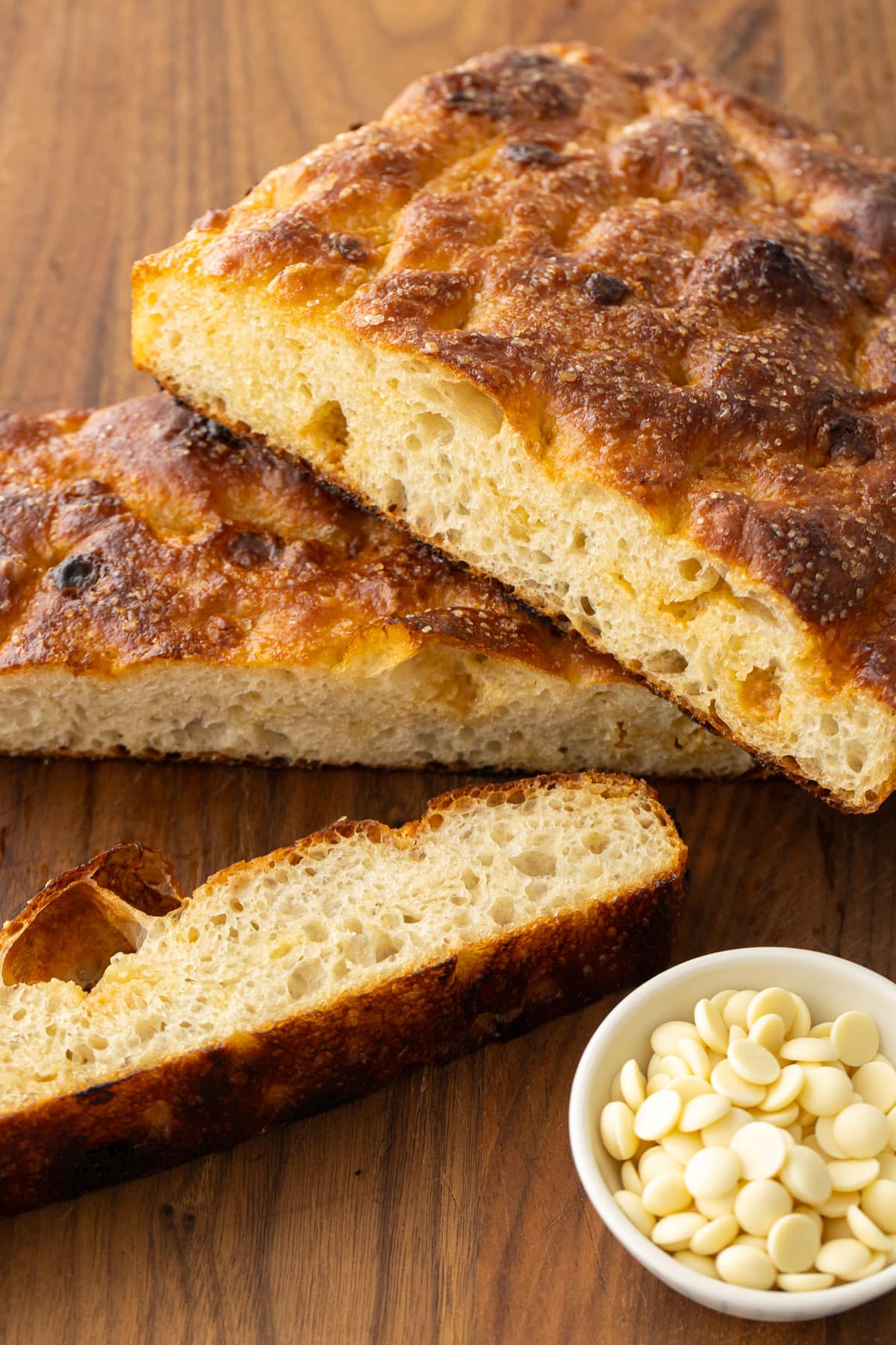 Slices of White Chocolate Focaccia Bread on a wooden cutting board with a bowl of white chocolate pieces on the side.