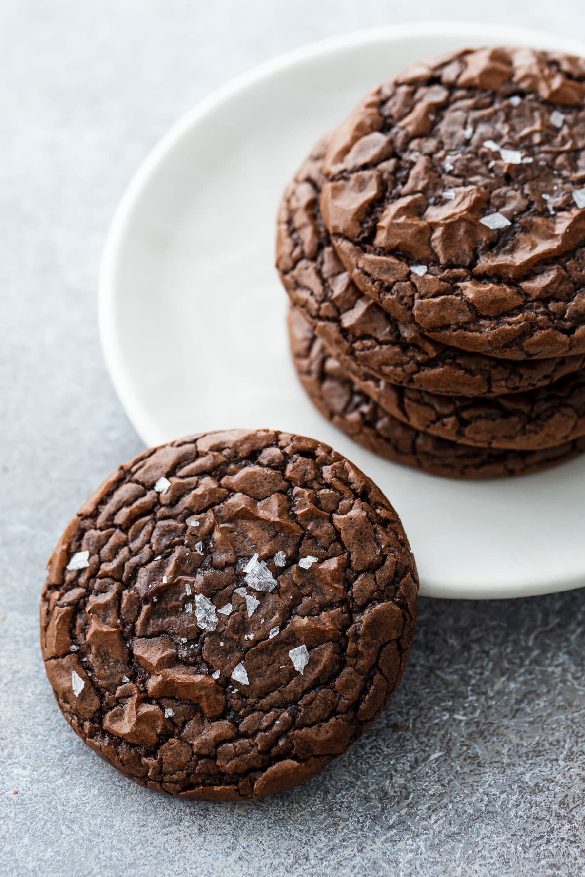 Stack of Chewy Crinkle-Top Brownie Cookies on a plate with one cookie propped on the rim to show the shiny, crackly top texture.