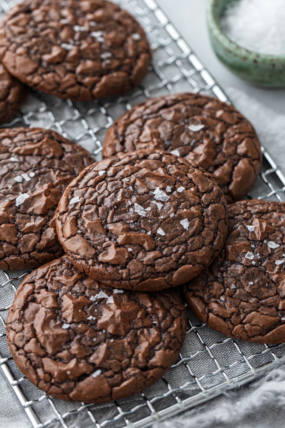 Chewy Crinkle-Top Brownie Cookies arranged haphazardly on a wire rack, with bowl of sea salt in the background.