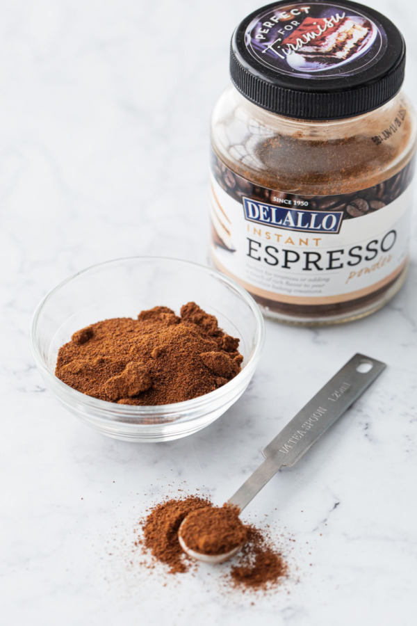 Jar of instant espresso powder on a marble background with a bowl and spoon full of espresso powder on the side.