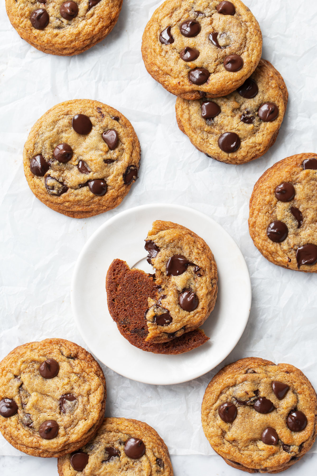 Overhead, scattered Brown Butter Banana Bread Chocolate Chip Cookies on a white parchment background, one cookie broken in half to show the texture.