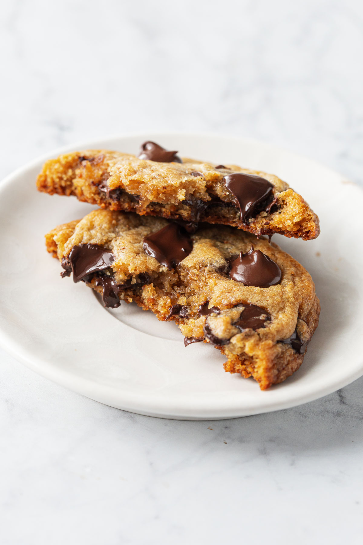 Closeup of a Brown Butter Banana Bread Chocolate Chip Cookie on a white plate, broken in half to show the melty chocolate and gooey texture inside.