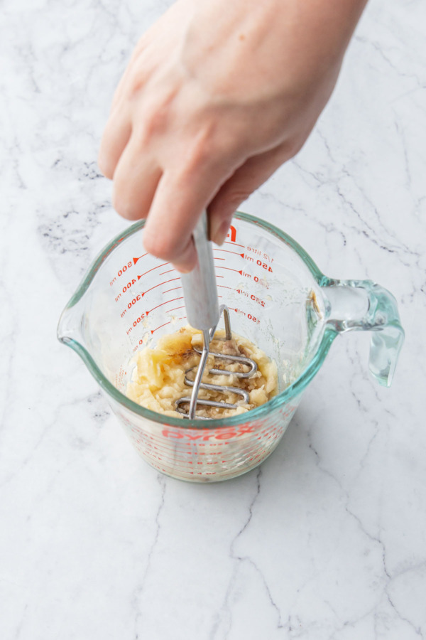 Mashing a very ripe banana in a glass measuring cup with a small potato masher.