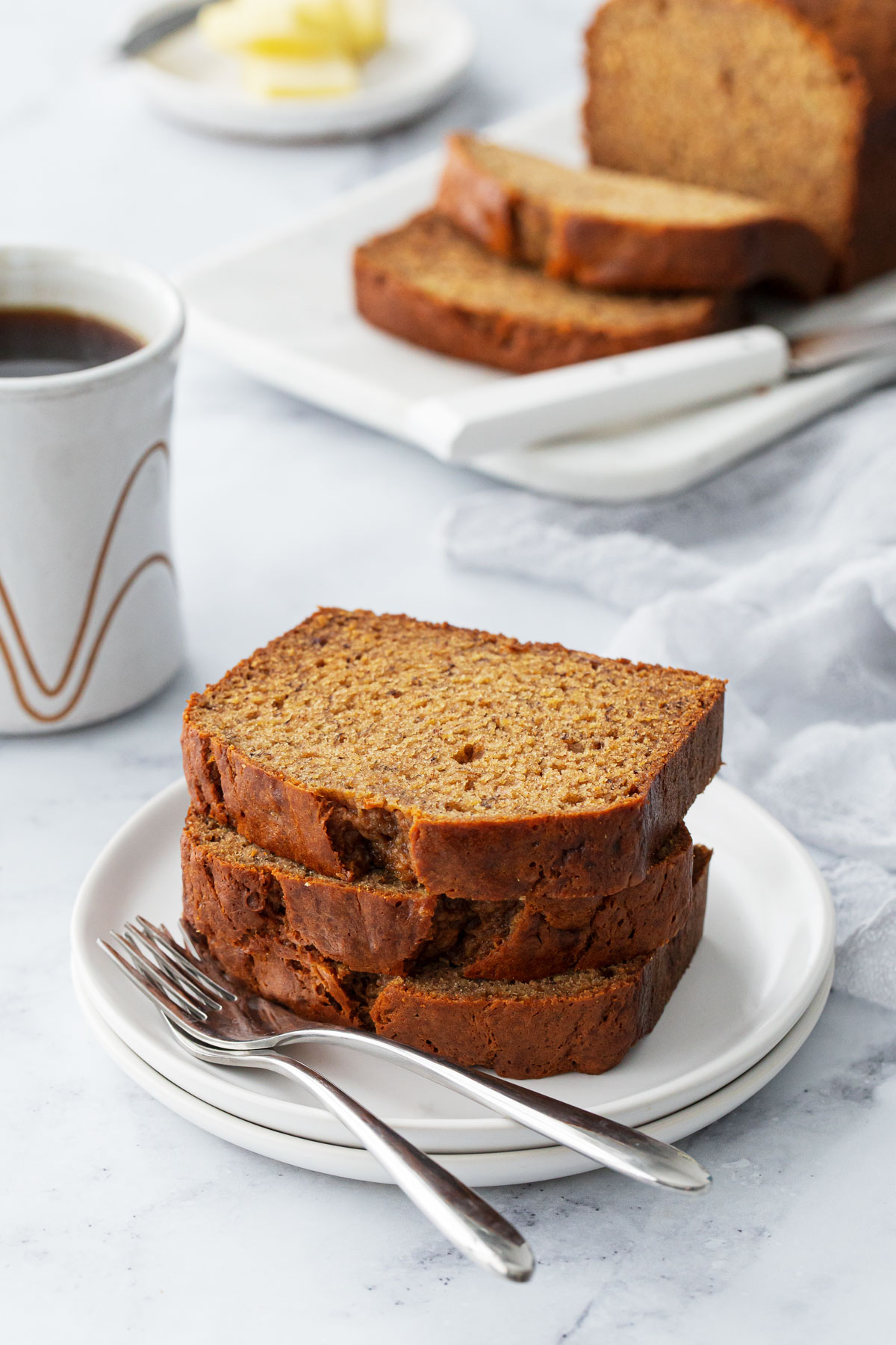 Stack of three slices of Brown Butter Banana Bread on a white plate, with coffee cup and sliced loaf in the background.
