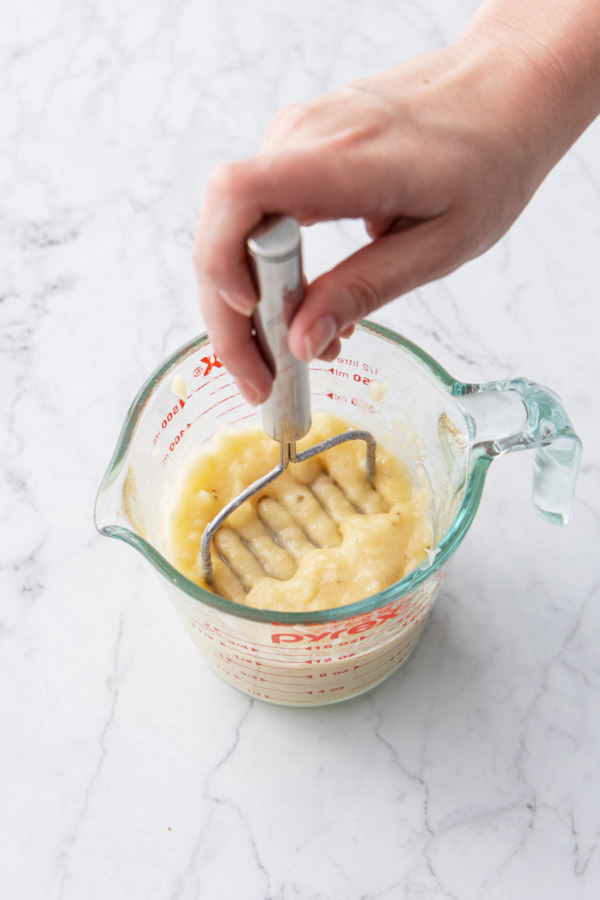 Mashing ripe bananas with a small wire potato masher in a 2-cup glass measuring cup.