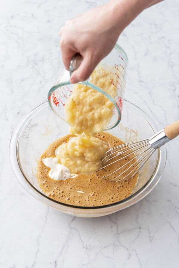 Pouring mashed banana into glass mixing bowl along with egg/sugar mixture and sour cream.