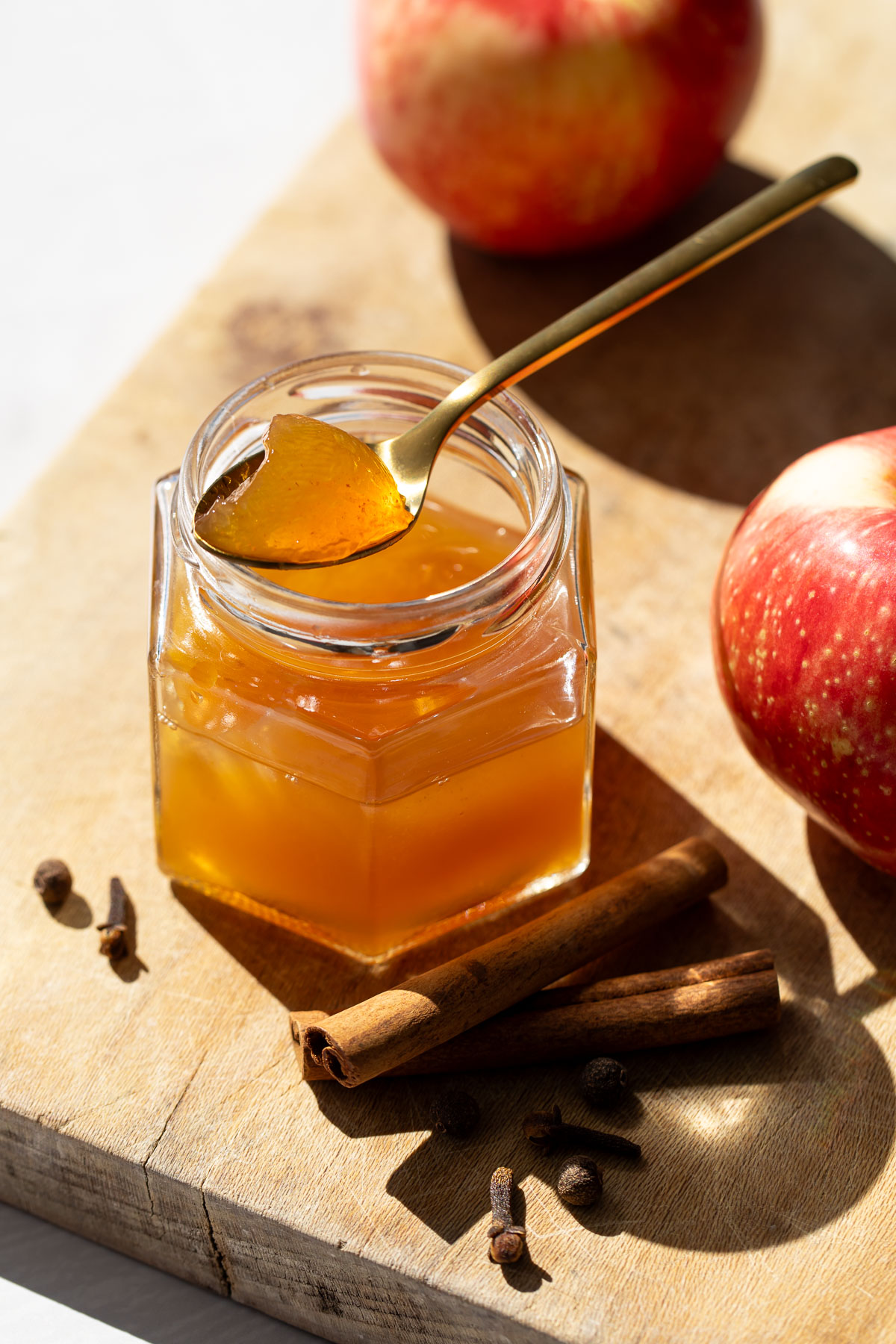 Jar of Spiced Apple Cider Jelly on a wooden board, a spoonfull of jelly resting on the rim of the jar, with fresh apples and whole spices.