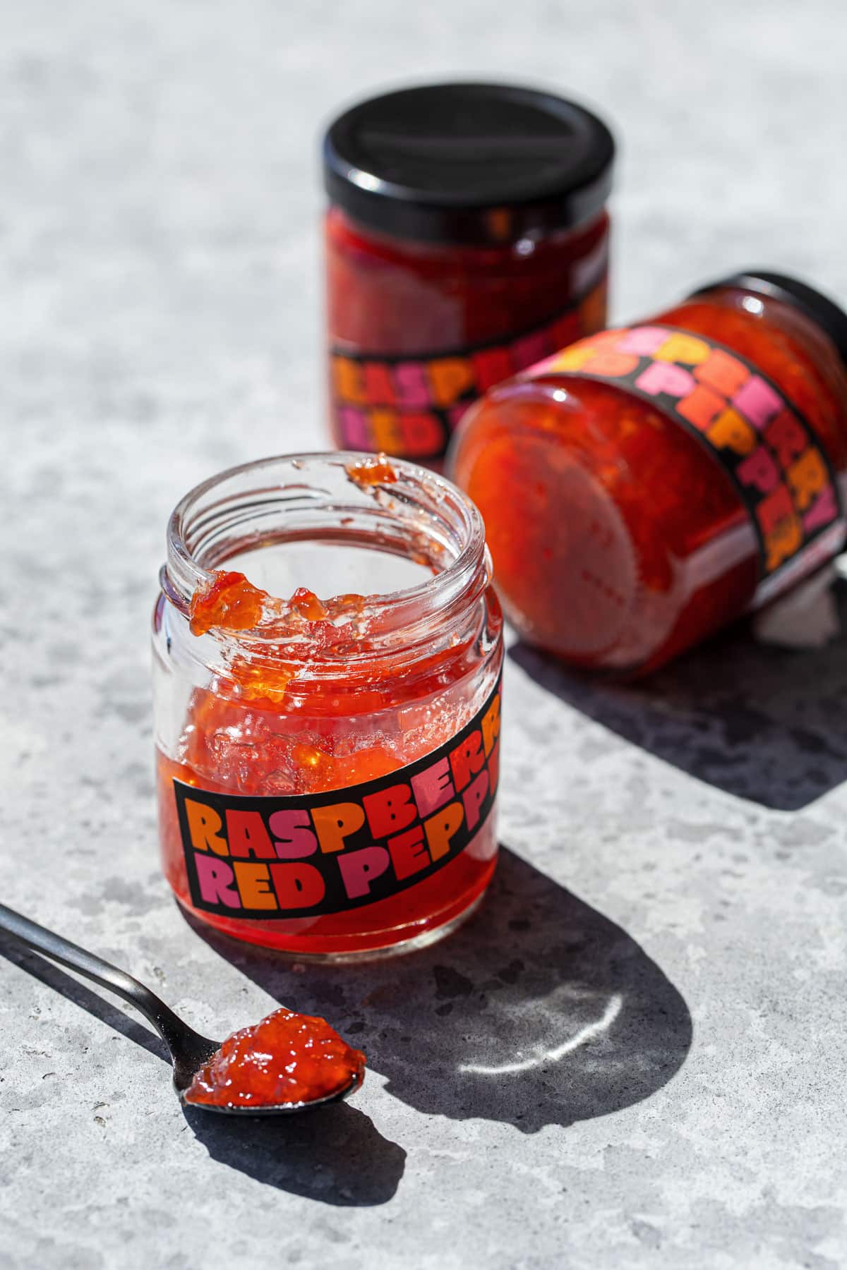 Three jars of Raspberry Red Pepper Jelly, one open and a spoonful sitting in the foreground to show the texture of the jelly.