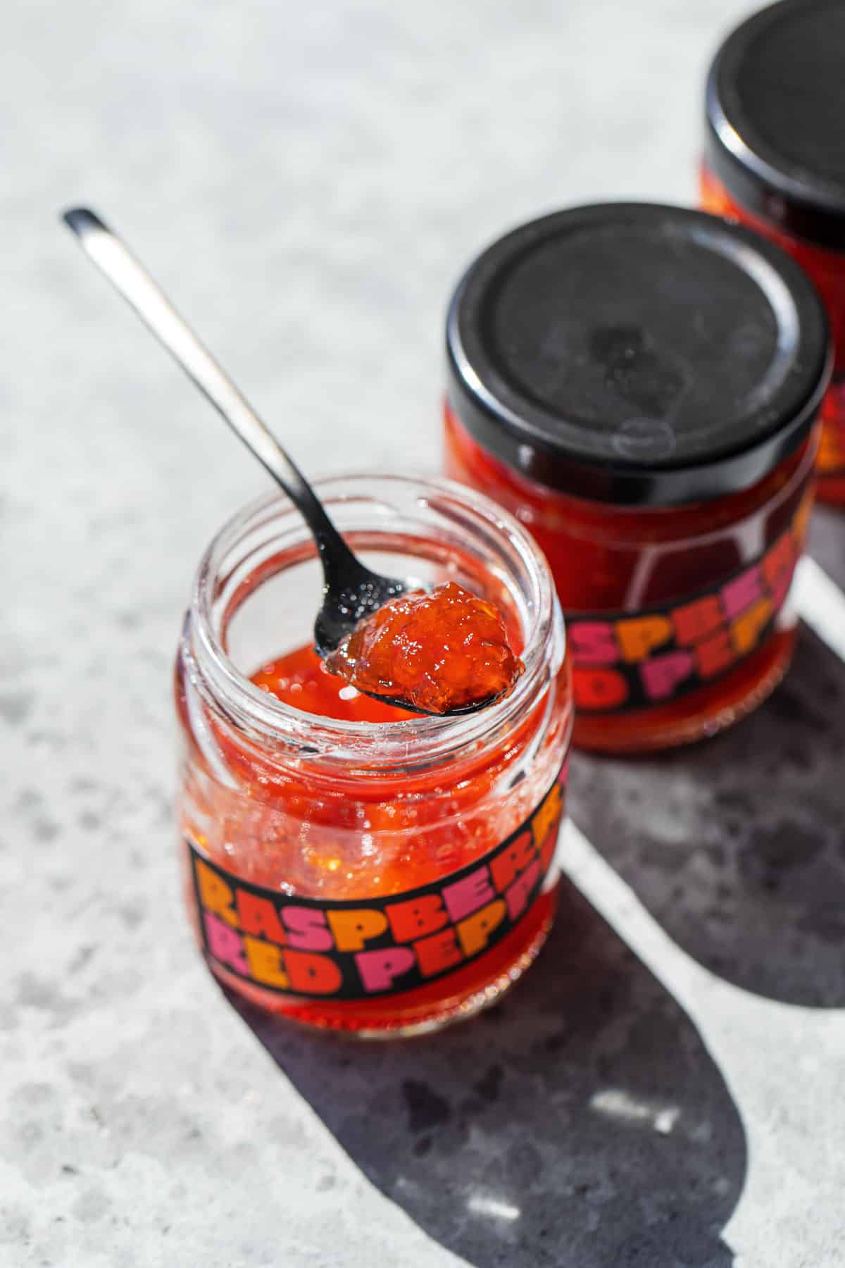Jars of Raspberry Red Pepper Jelly with custom labels, one jar open with a spoonful resting on top to show texture.
