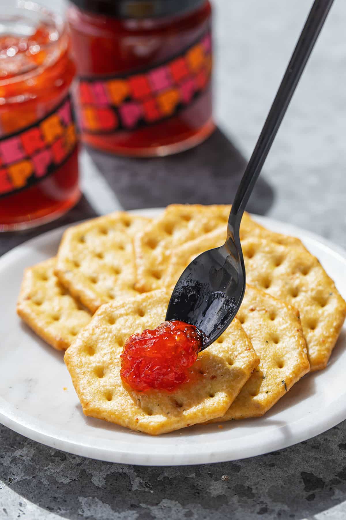 Spoon dropping a dollop of Raspberry Red Pepper Jelly onto a cracker sitting on a plate, jars of jelly in the background.